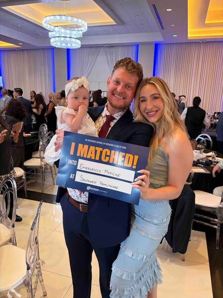 Andrew McLain with his wife and baby, holding an "I matched!" sign