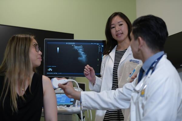 A medical student teaches another student how to use an ultrasound device while a standardized patient looks on.