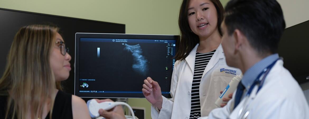 A medical student teaches another student how to use an ultrasound device while a standardized patient looks on.